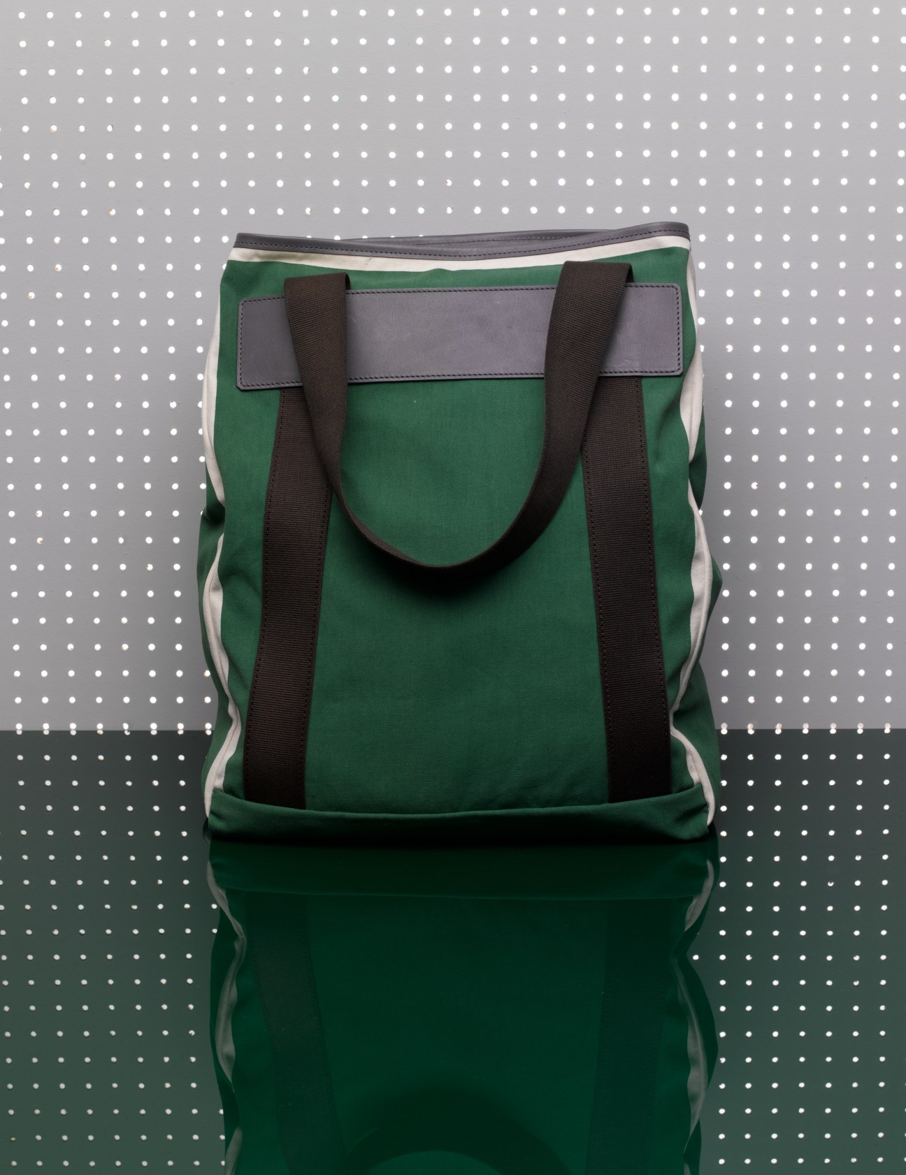 Green canvas tote bag with black handles and gray leather trim against polka dot background.