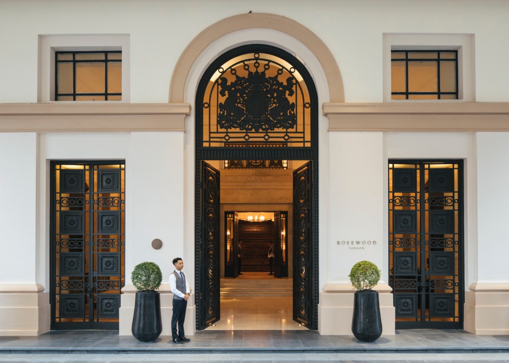 Hotel staff member standing at entrance of Rosewood Yangon with ornate archway and decorative planters