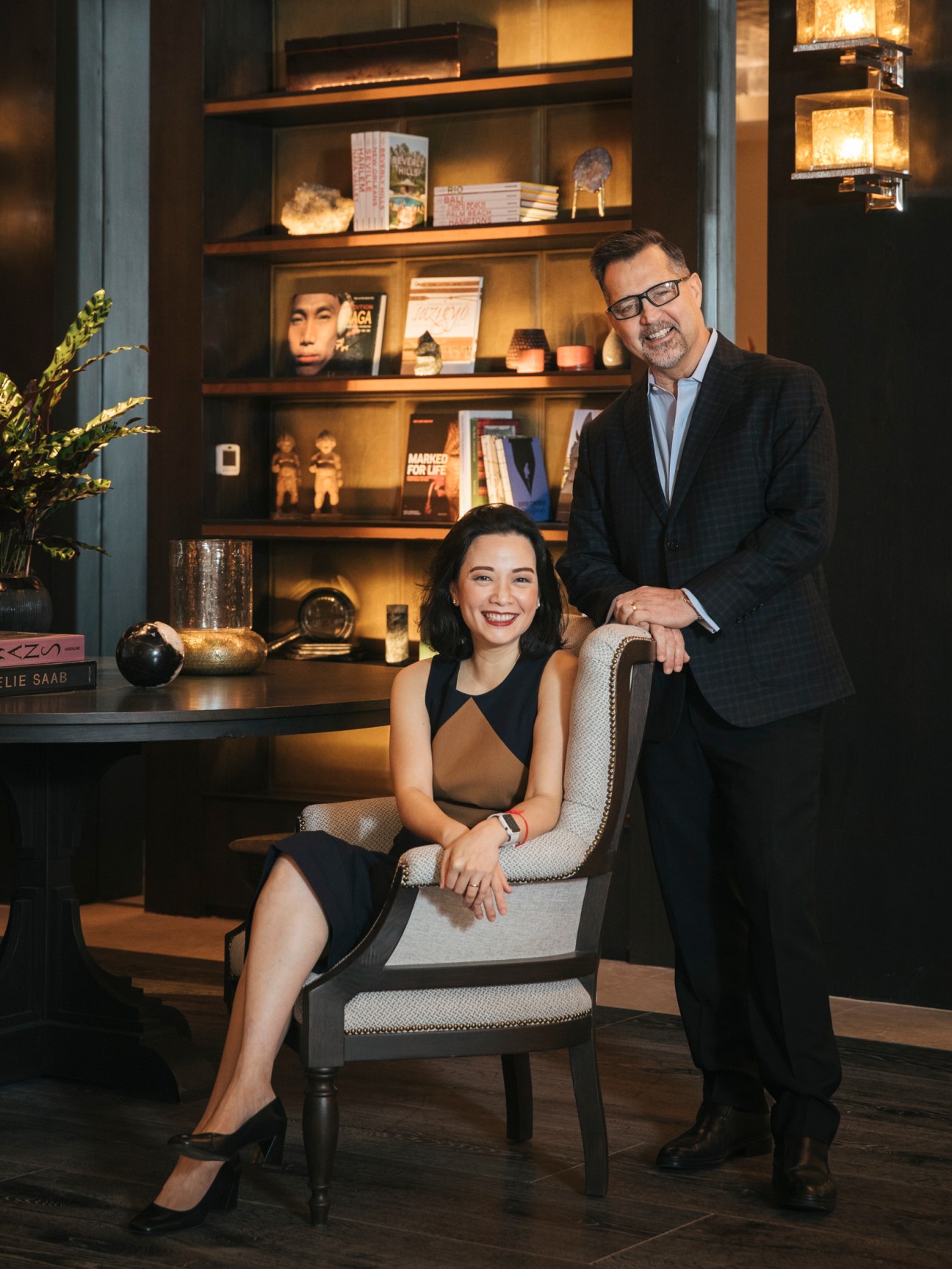 Supalak Foong seated in chair with Ed Brea standing beside her in upscale hotel interior with bookshelves