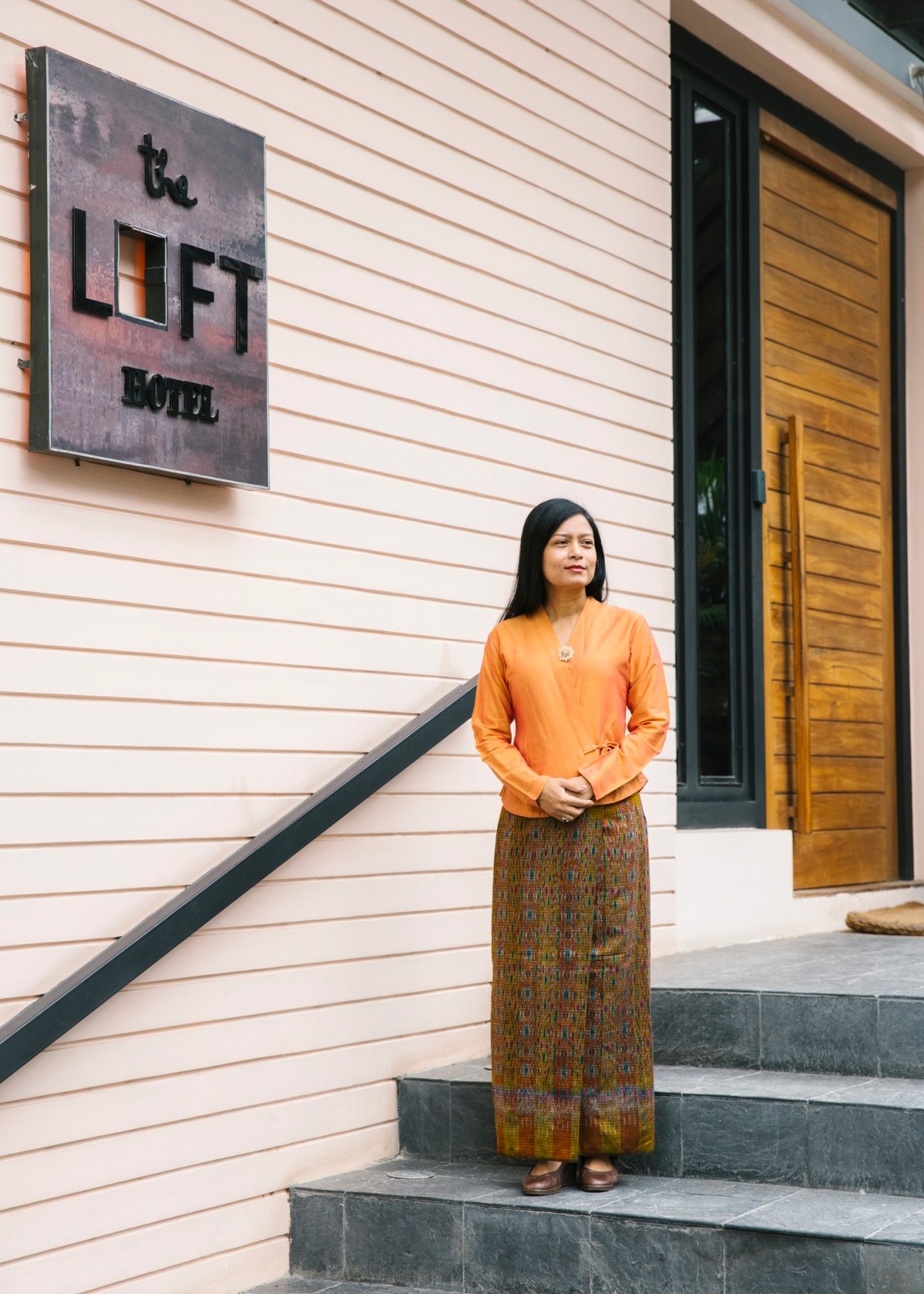 Sanda Win stands on steps outside The Loft Hotel wearing an orange top and patterned skirt