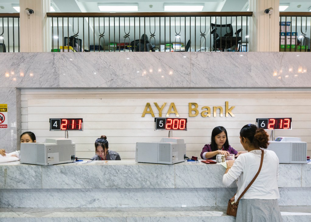 Bank tellers serving customers at marble counter with digital displays showing numbers 4, 5, and 6