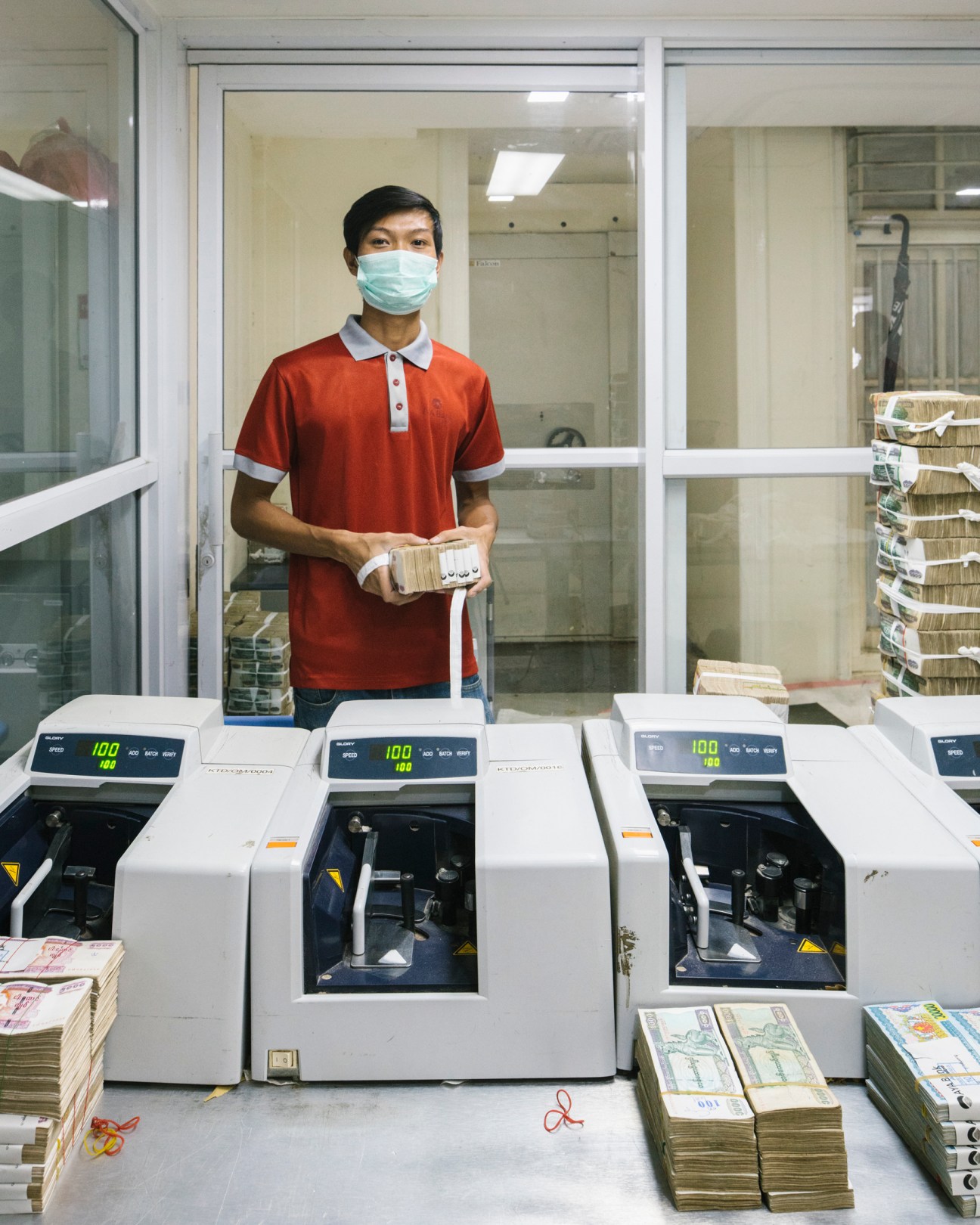 Bank employee in red polo shirt and face mask counting stacks of currency notes at Aya Bank in Yangon