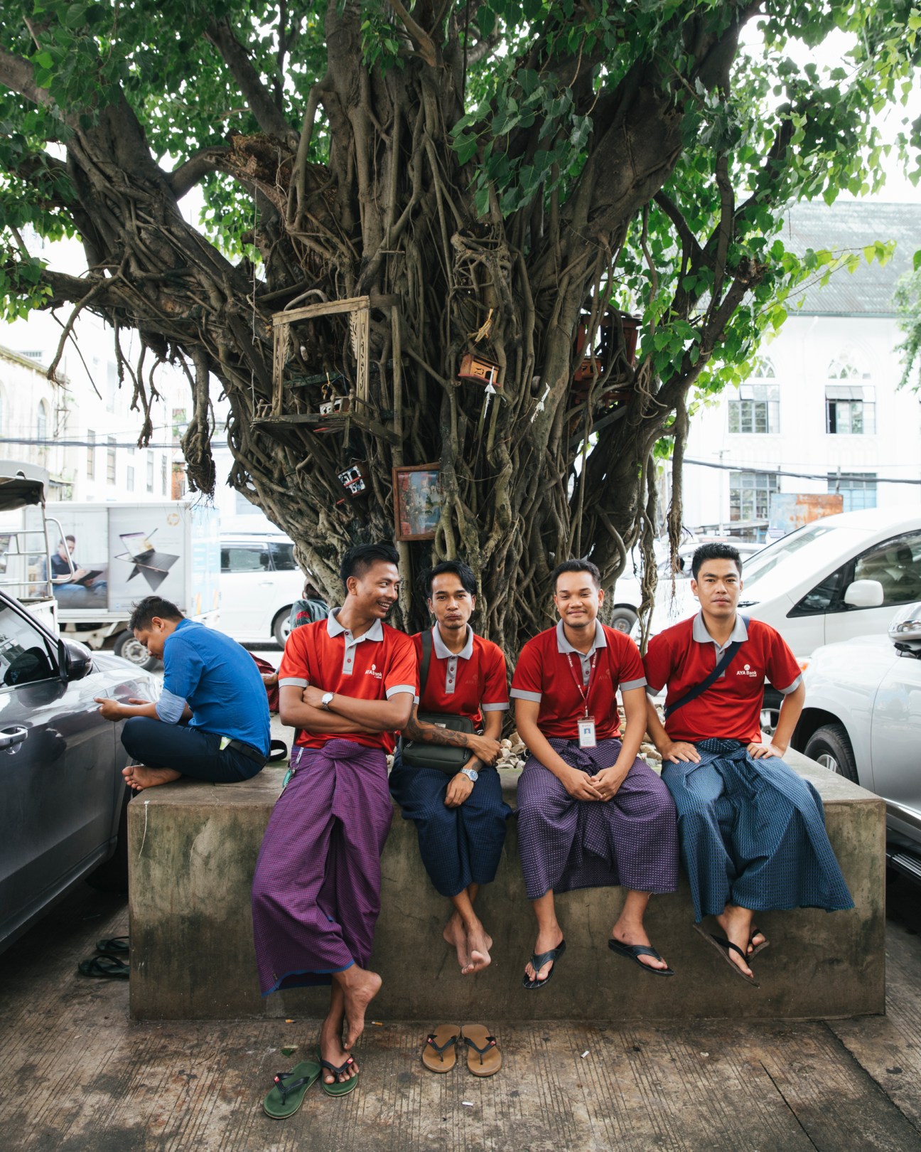 Four men in red polo shirts and traditional lungis sitting on concrete ledge under large tree