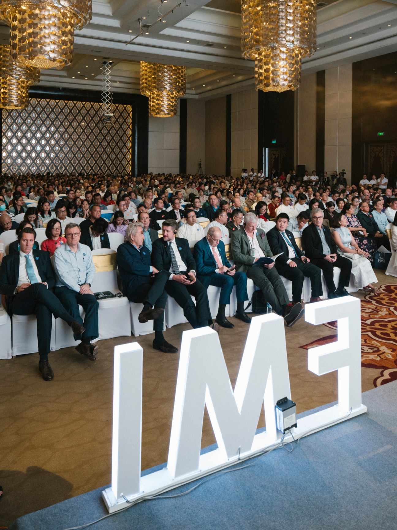 Large conference hall filled with hundreds of attendees seated in white chairs at corporate meeting