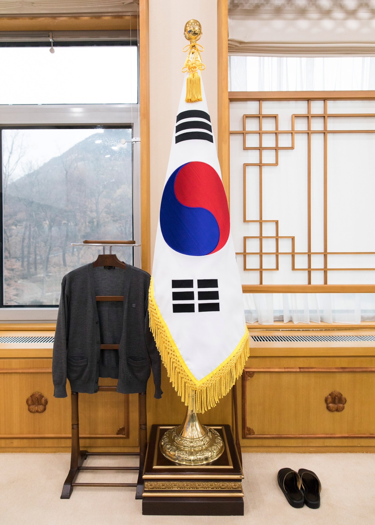 South Korean flag with gold tassel displayed in an office interior with wooden paneling and windows, grey cardigan on clothes rack nearby.