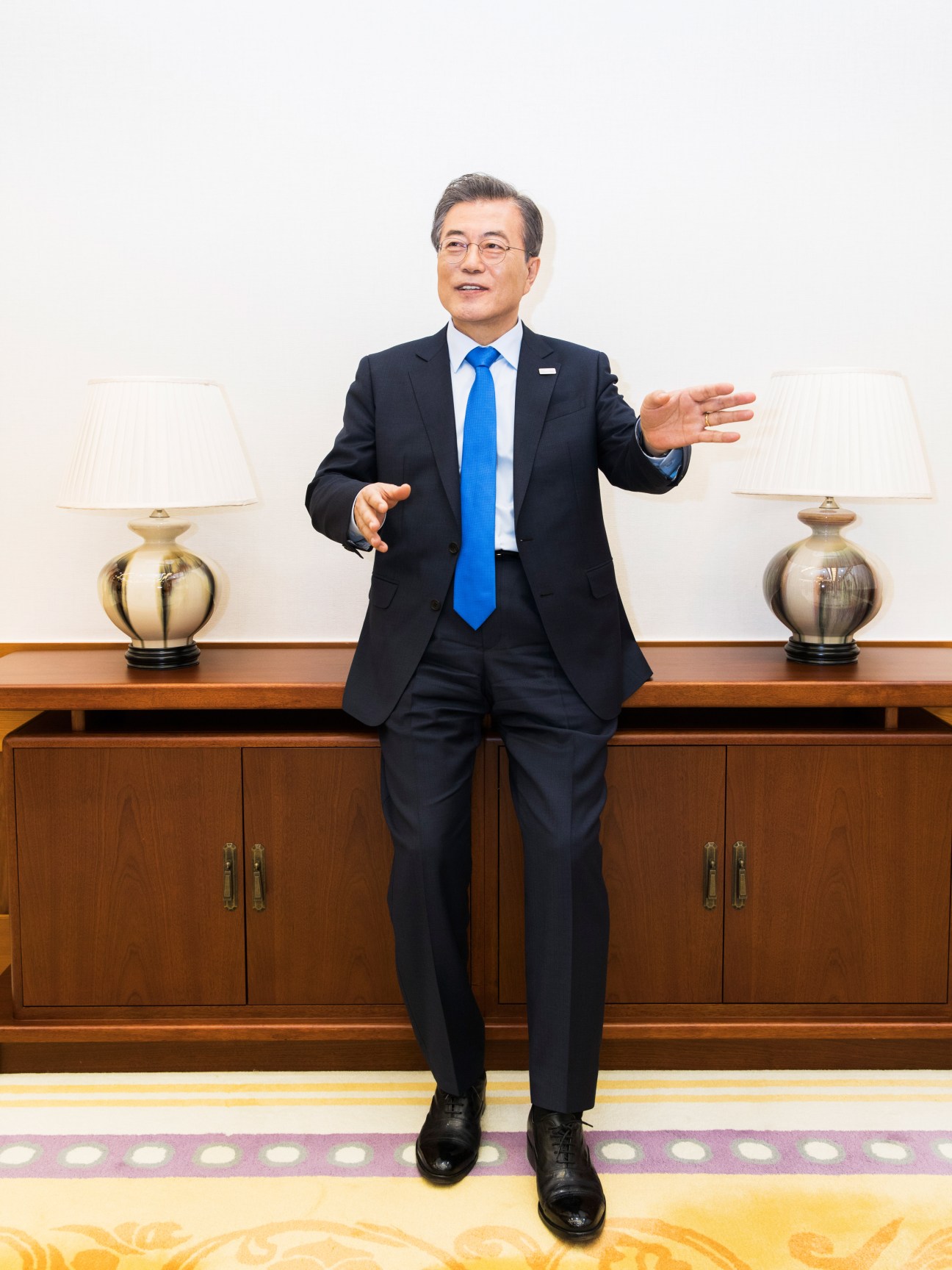 President Moon Jae-in stands in a formal pose wearing a dark suit and blue tie in an elegant room with gold lamps and wooden cabinetry.