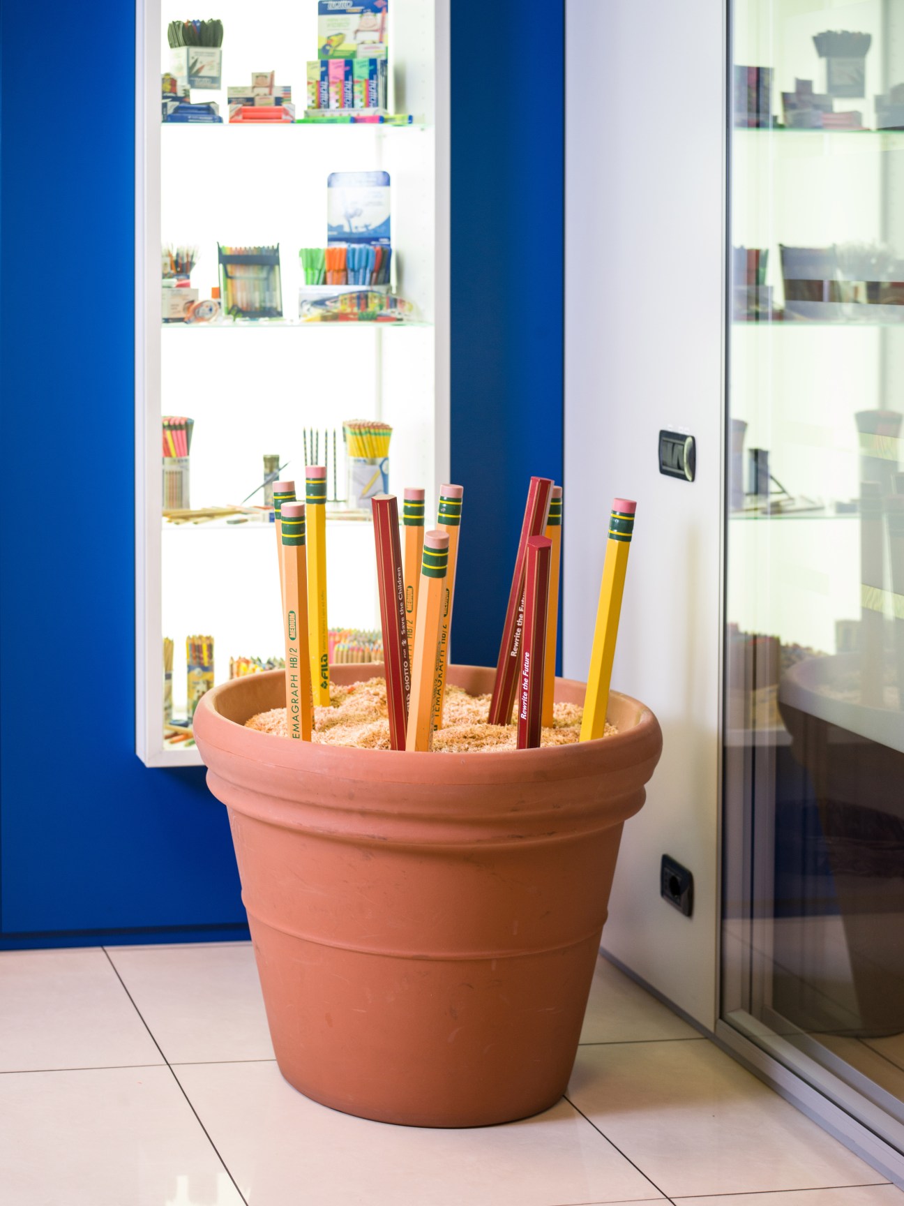 Terra cotta pot filled with pencils and erasers of varying sizes and colors against blue and white wall shelving.