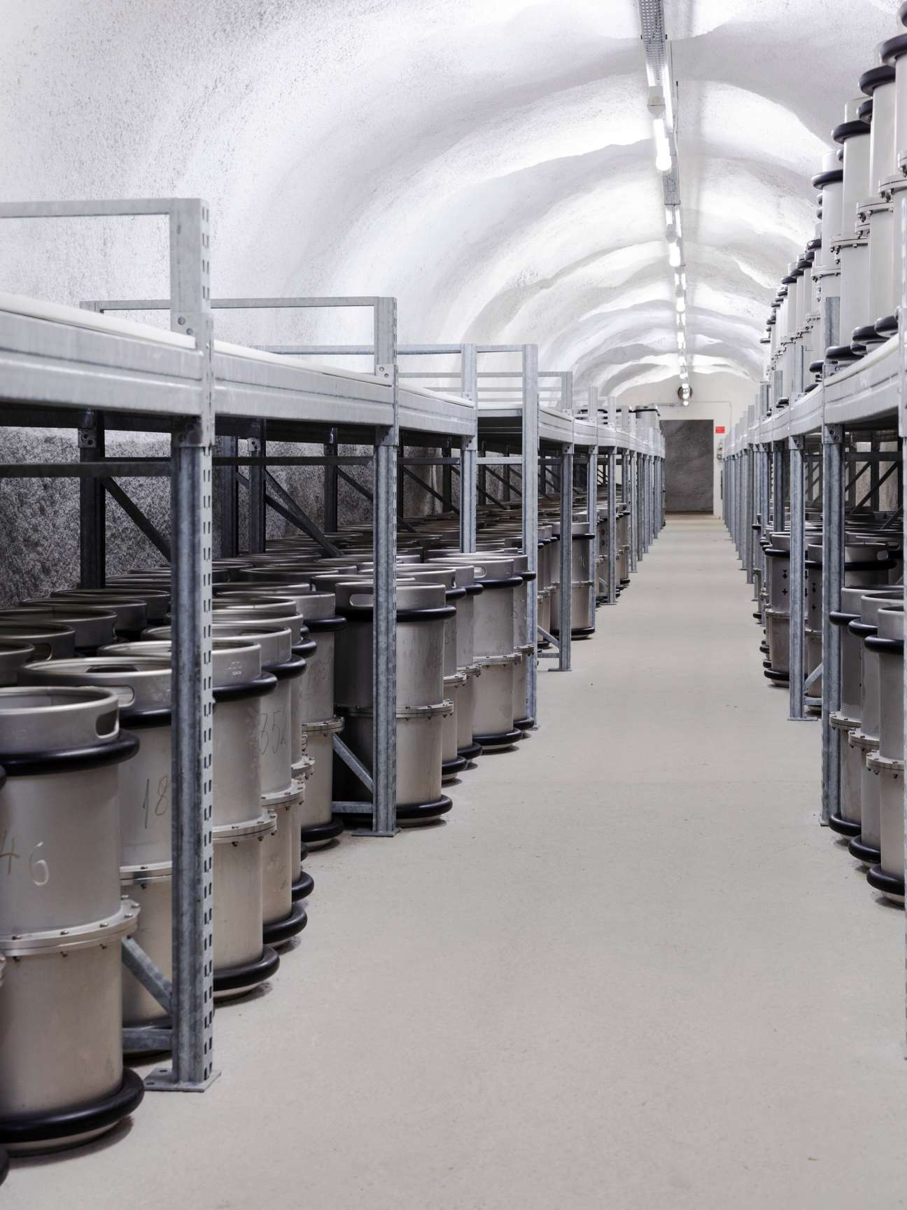 Metal cylindrical microfilm storage barrels arranged in rows inside an underground tunnel with arched ceiling and fluorescent lighting.