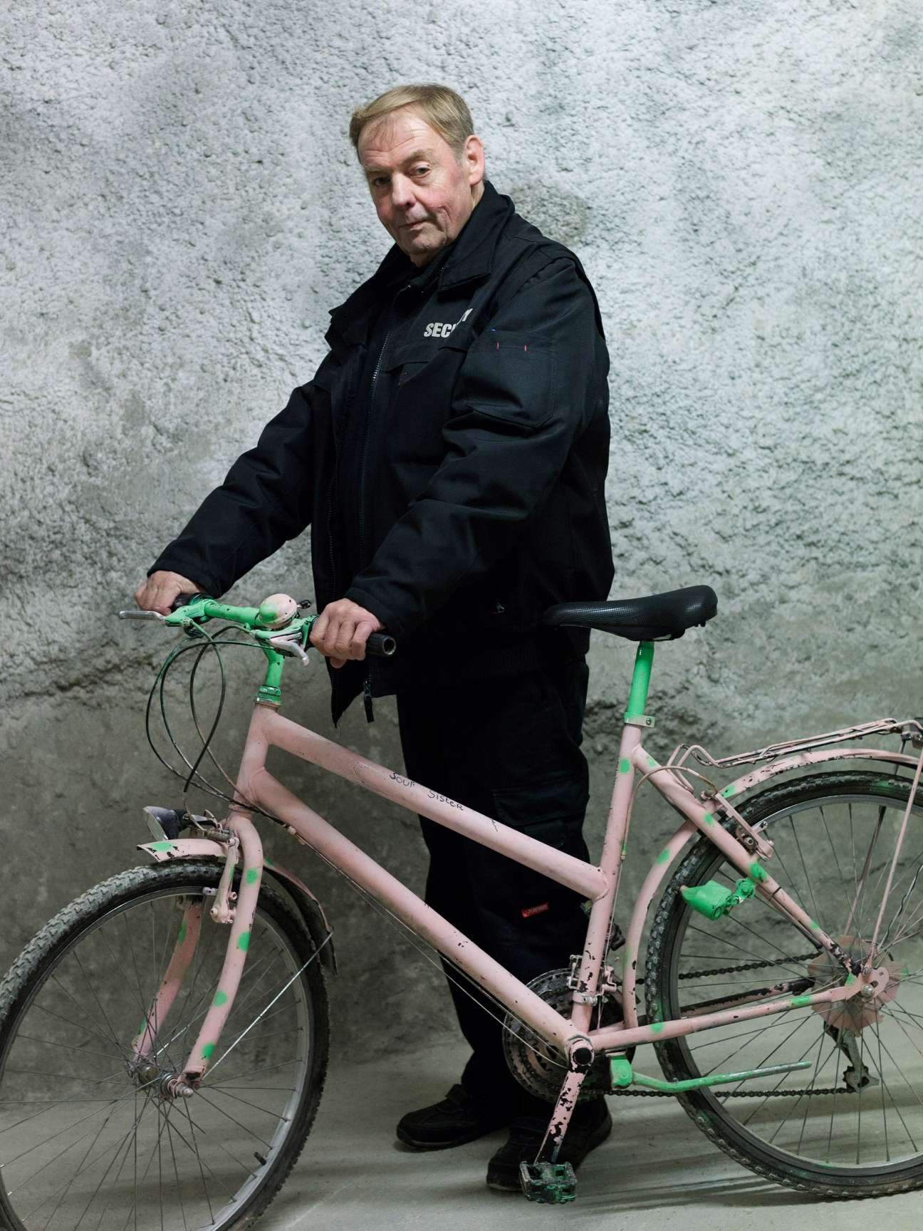 Michael Peldt, a security employee, poses with an old pink and green bicycle against a concrete wall.