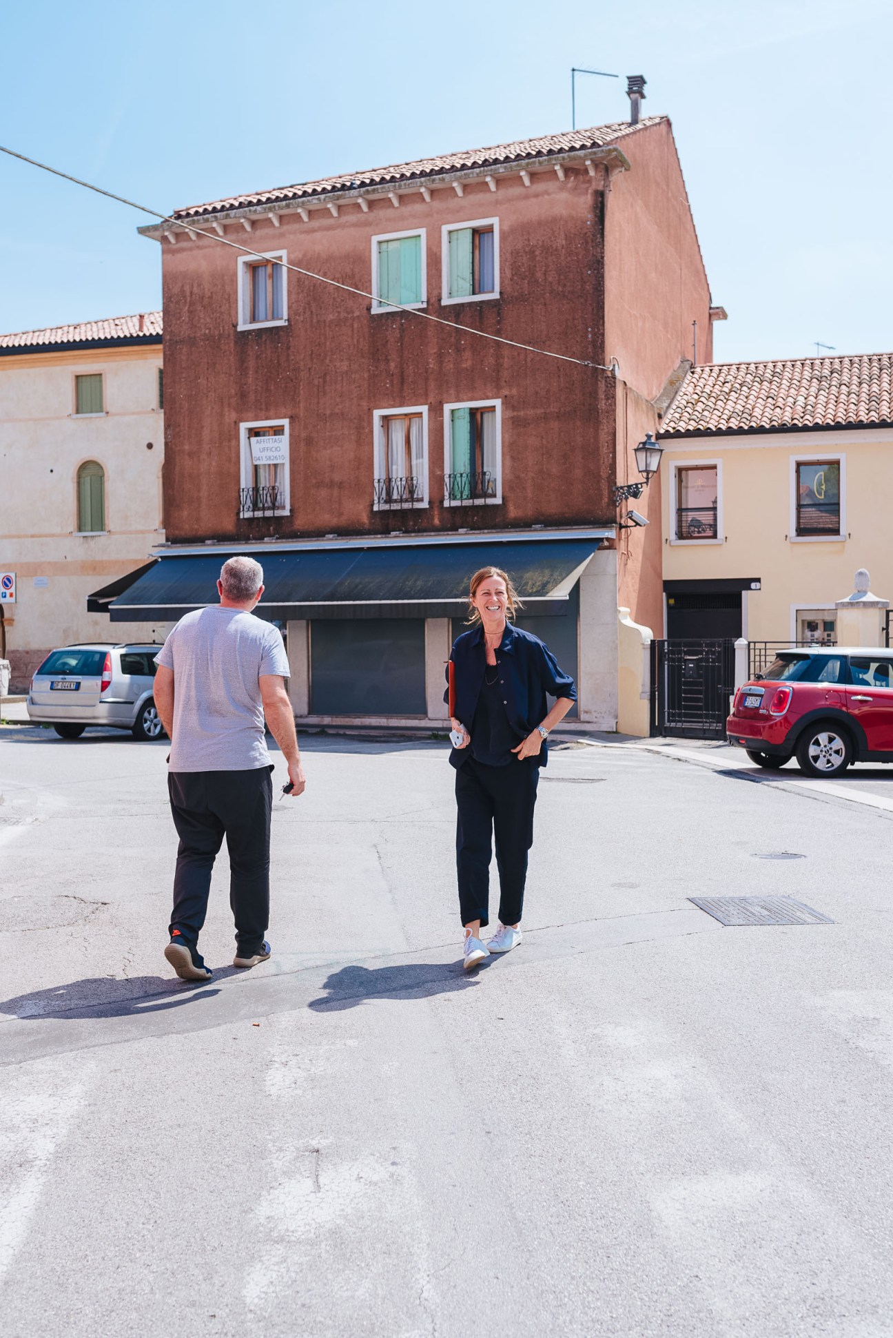 Massimo Pigozzo and Francesca Zara walking through a sun-drenched square in Mirano with a terracotta building behind them.