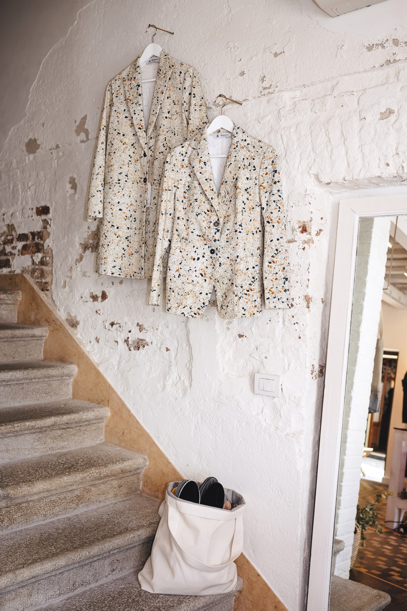 Terrazzo-patterned blazers hanging on a white studio wall above a canvas tote bag.