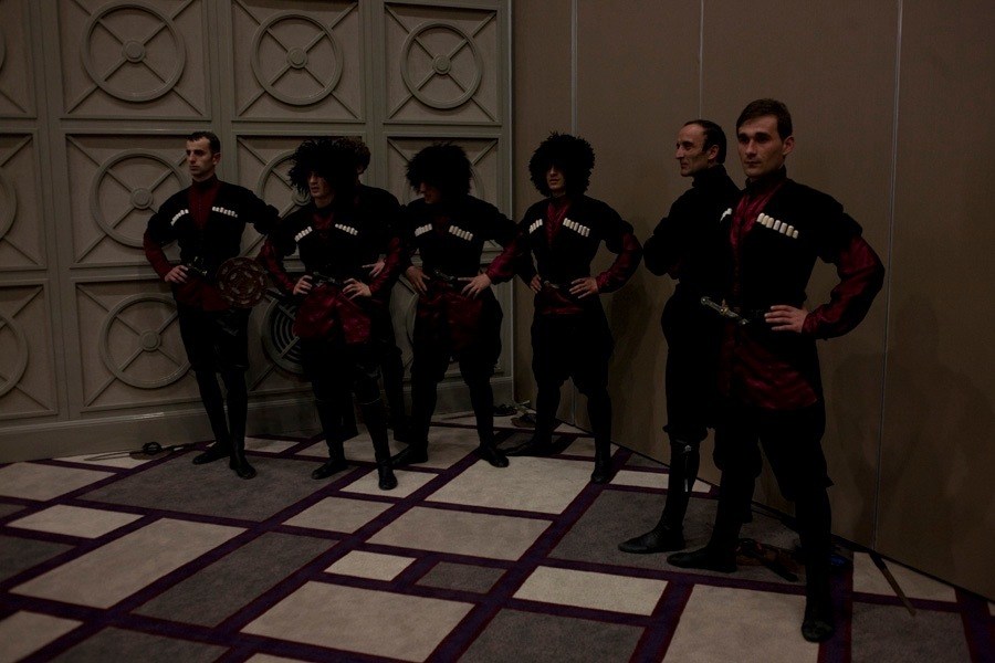 Six performers in traditional Georgian black and red costumes with decorative medallions stand posing against a paneled wall.