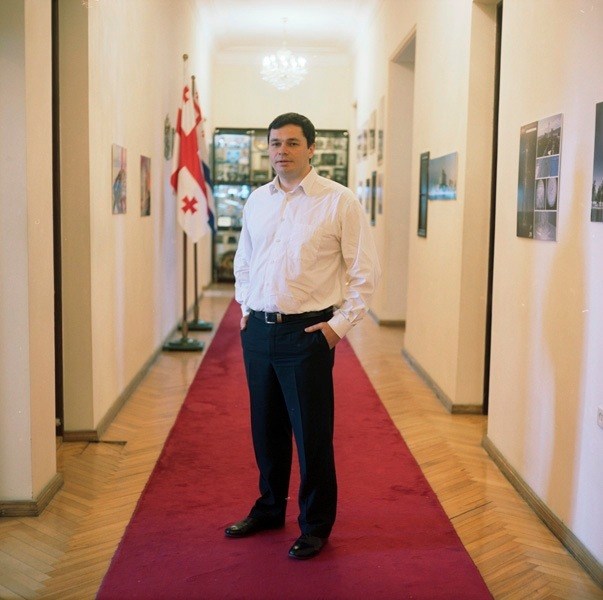 Man in white shirt and dark trousers standing on red carpet in hallway with Georgian flag visible