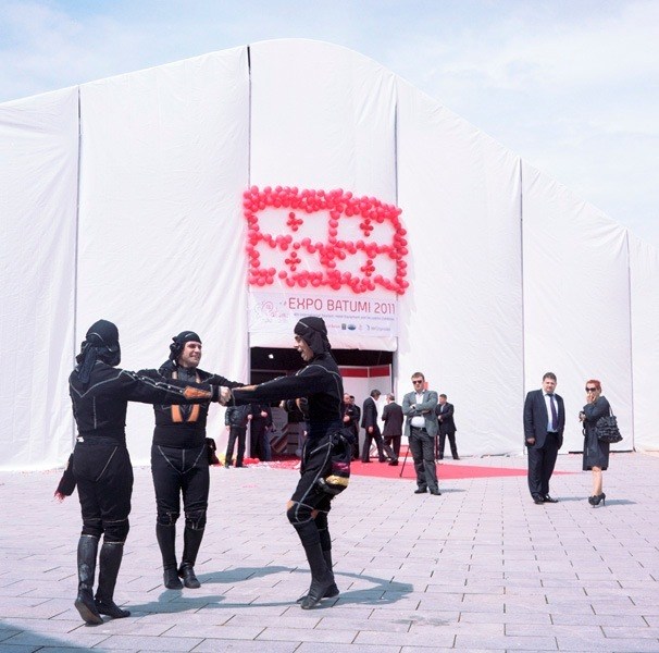 Traditional dancers in black costumes perform outside a large white tent structure with pink lettering for Expo Batumi 2011.