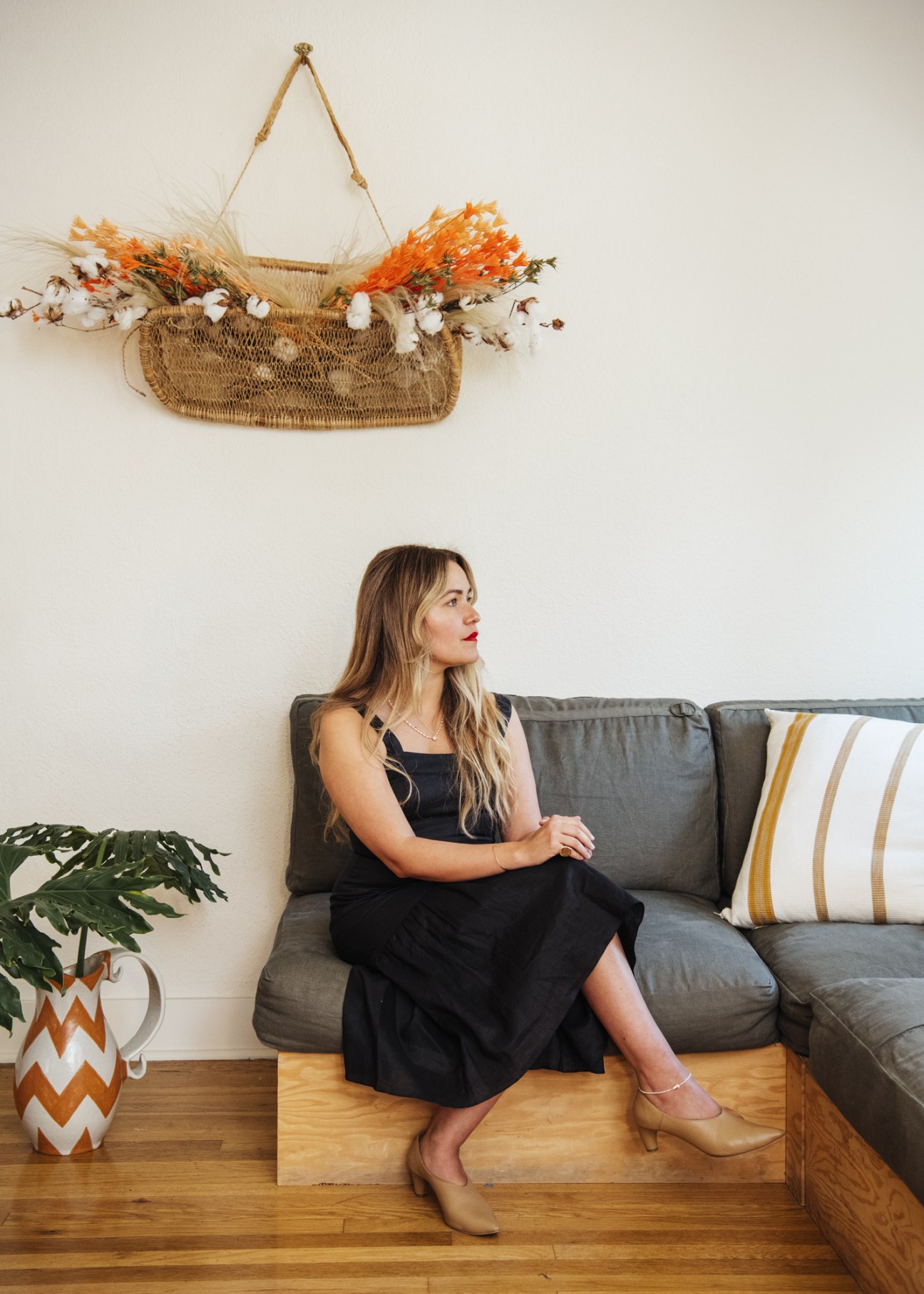Woman sitting on a gray sofa wearing a black dress, with a woven wall hanging and patterned ceramic vase visible behind her.