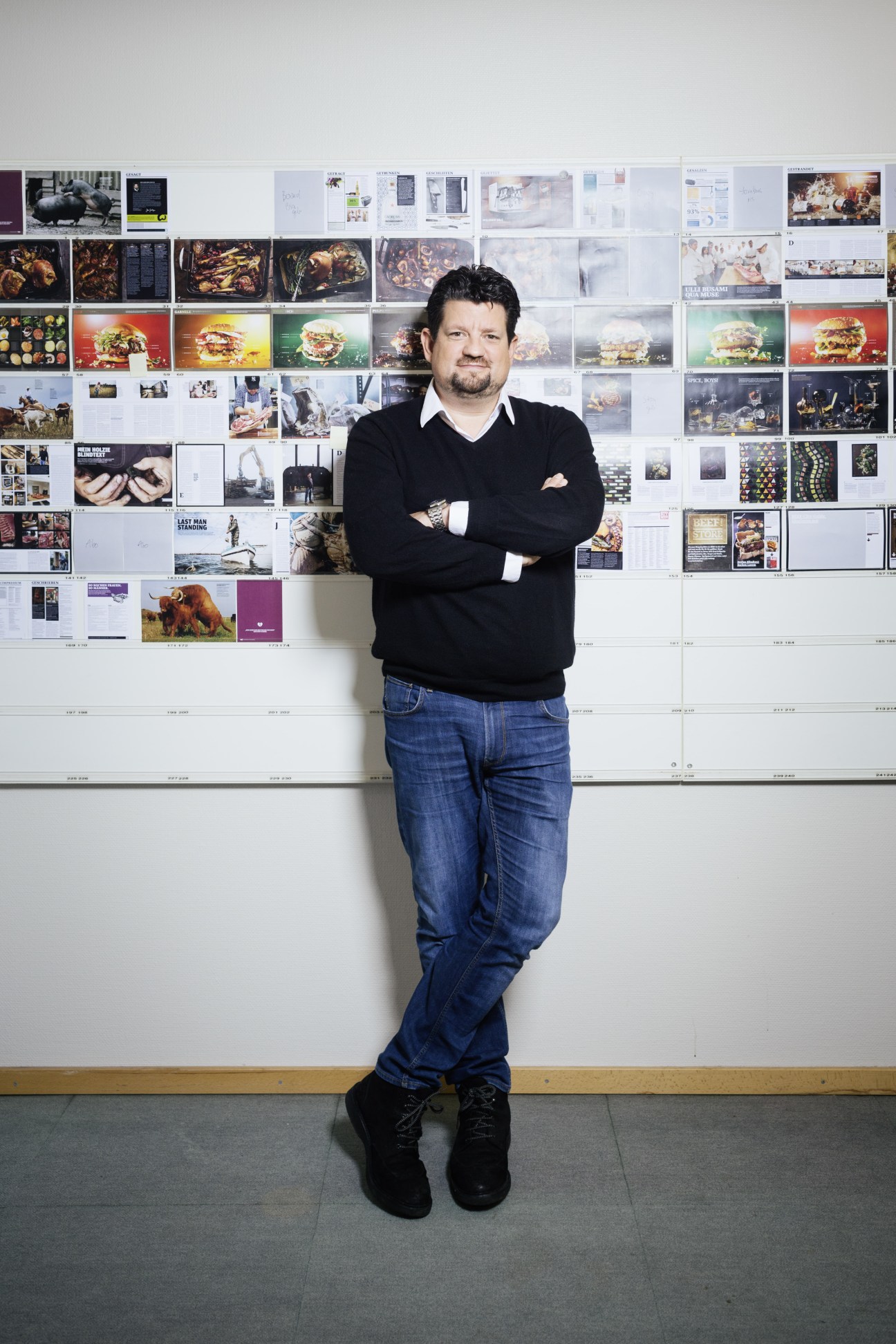 Jan Spielhagen, founder and editor of Beef! magazine, standing with arms crossed against a wall of magazine layouts and food photography.