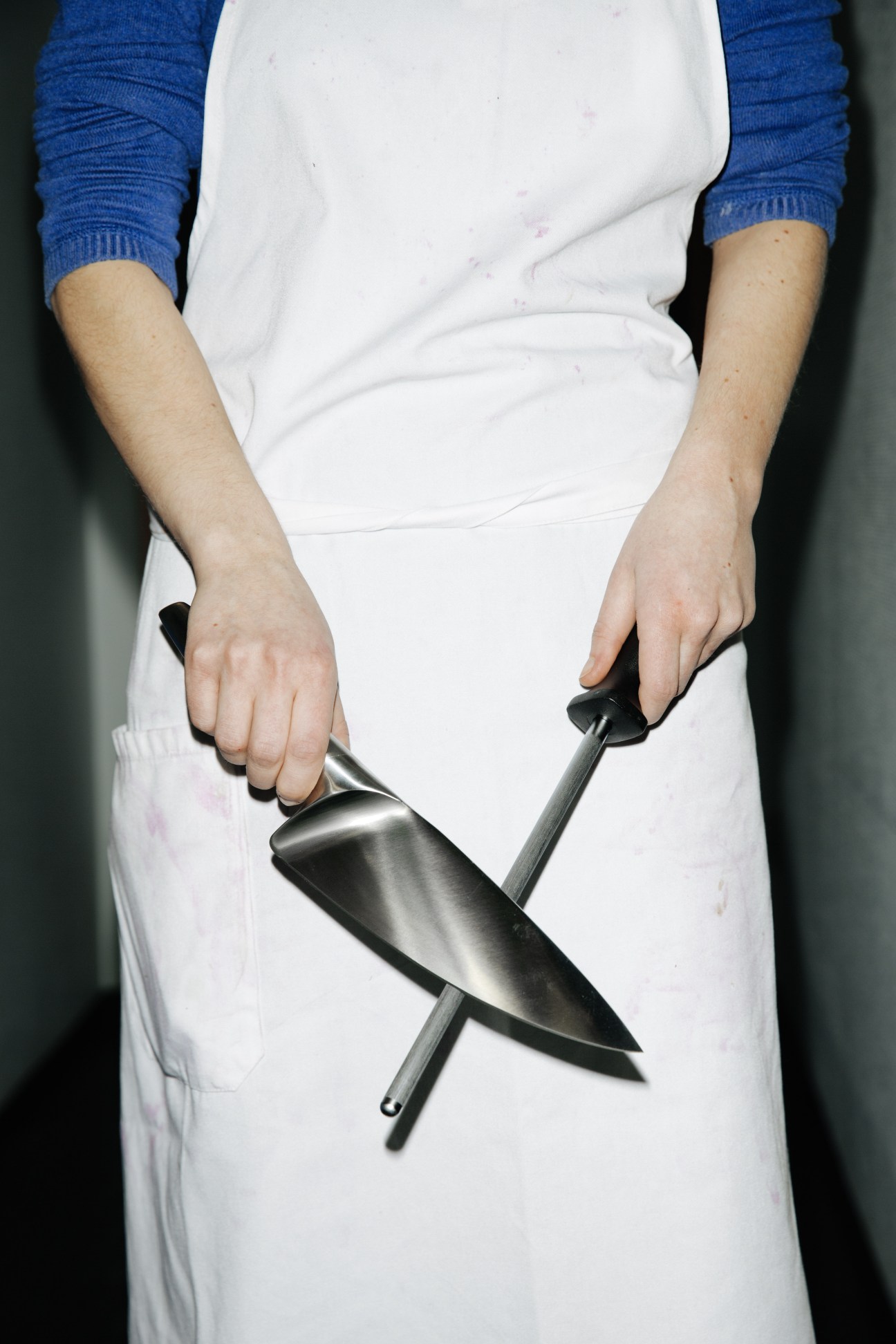 Person in white apron and blue sleeves holding a large kitchen knife and sharpener