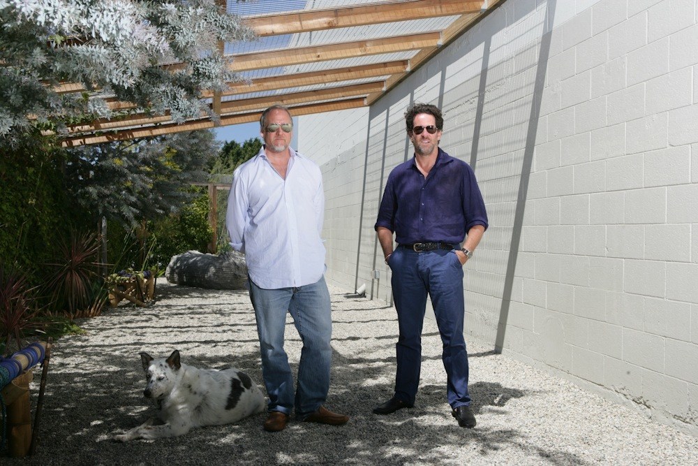 Two men stand in a sunlit courtyard with white walls and wooden beams; a dog rests in the foreground.