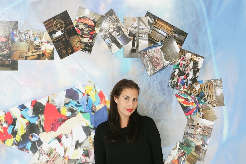 Lauri Firstenberg, director of LAXART, poses in front of a light blue wall with photographs and colorful collage artwork arranged in a circular pattern.