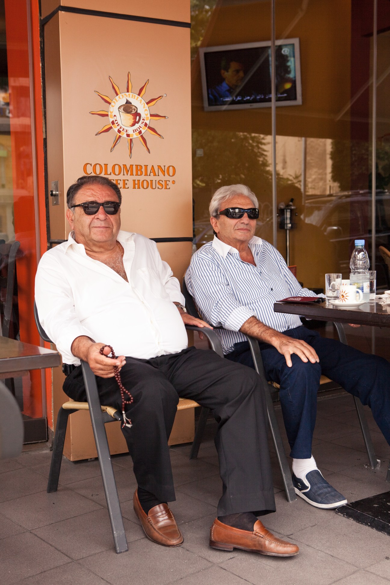 Two men wearing sunglasses sit outside Colombiano Coffee House, with branding visible on the window behind them.