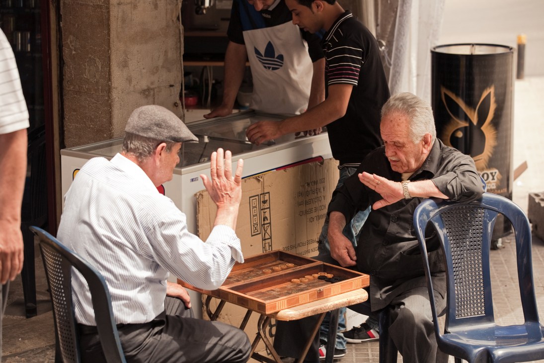 Two elderly men play backgammon on a street in Istanbul while a young vendor serves customers at a shop behind them.