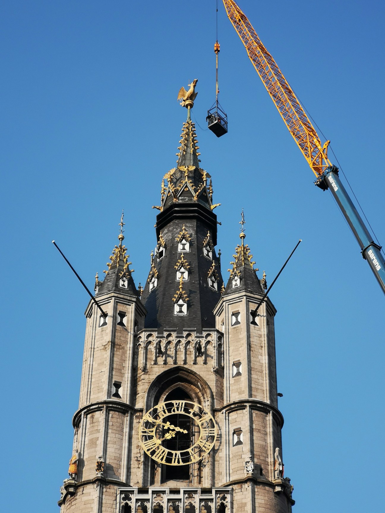 A gothic stone bell tower with ornate spires and a construction crane lifting materials during renovation work under clear blue sky.