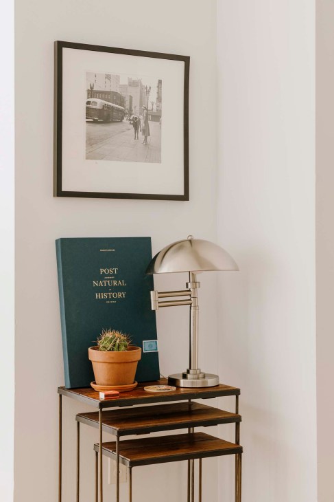 Framed black-and-white street photograph by Vivian Maier hanging above a wooden shelf display with a book, potted cactus, and desk lamp.