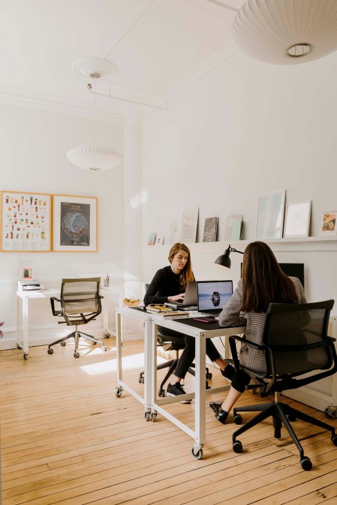 Two women work at a desk in a minimalist office with white walls displaying framed artwork and prints.