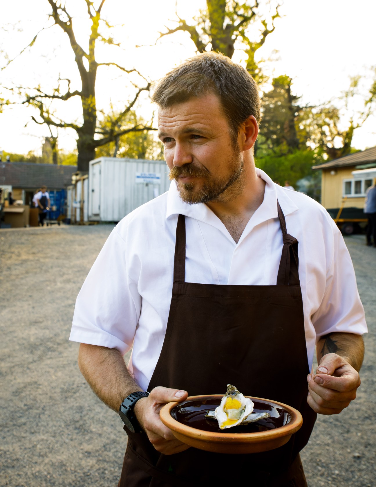 Billy White in white shirt and brown apron holds a ceramic bowl with an oyster outdoors