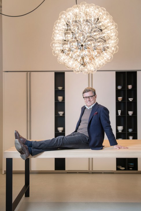 Roberto Gavazzi, head of Boffi, sits on a wooden table in a modern showroom with built-in shelving and a decorative crystal chandelier overhead.