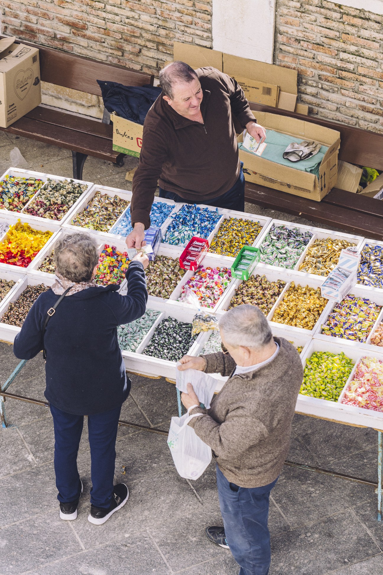 Man serving colorful wrapped candies to two customers at an outdoor market stall in Estepa.