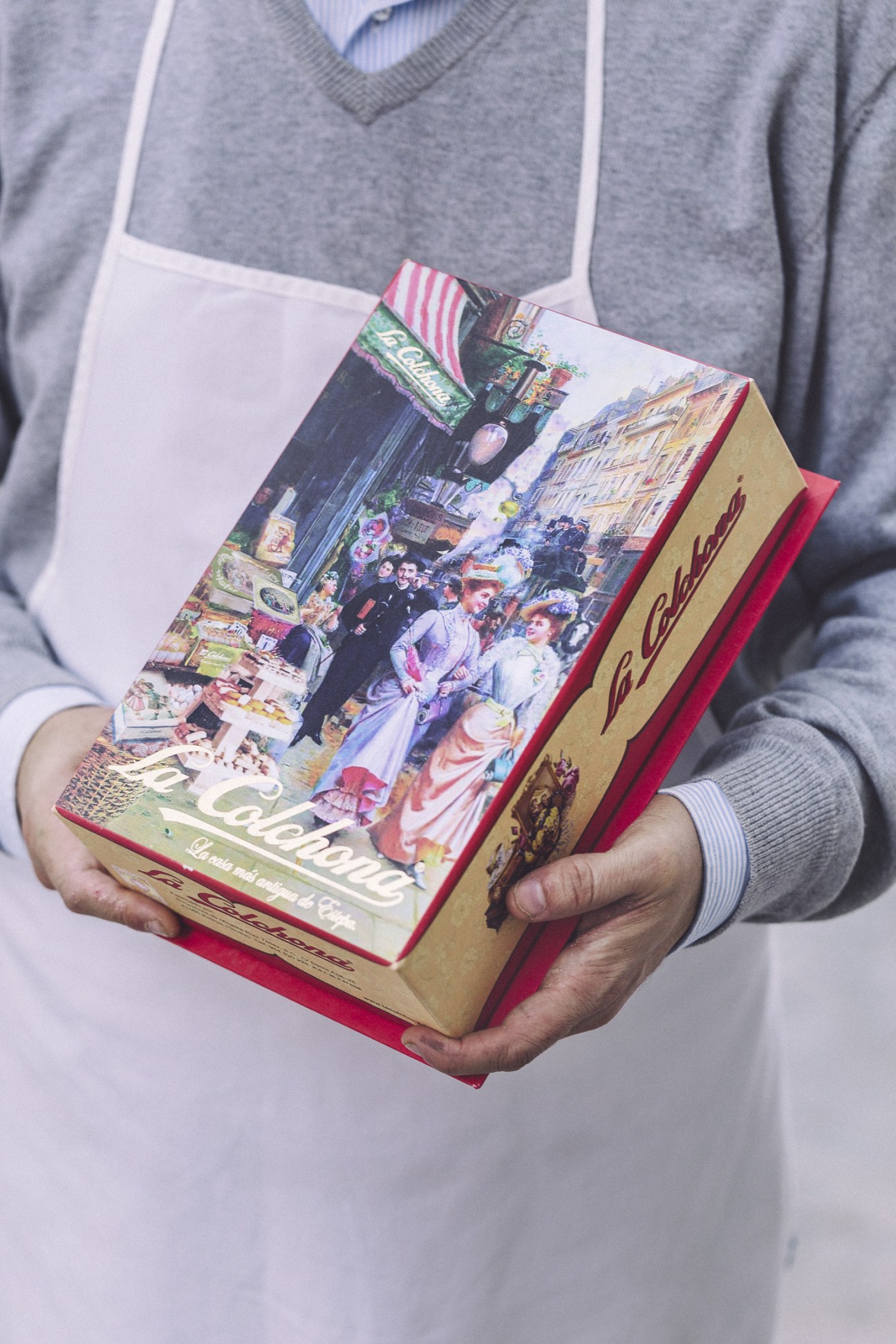 Hands holding a La Colchona box of mantecados with vintage-style packaging featuring a street scene illustration.