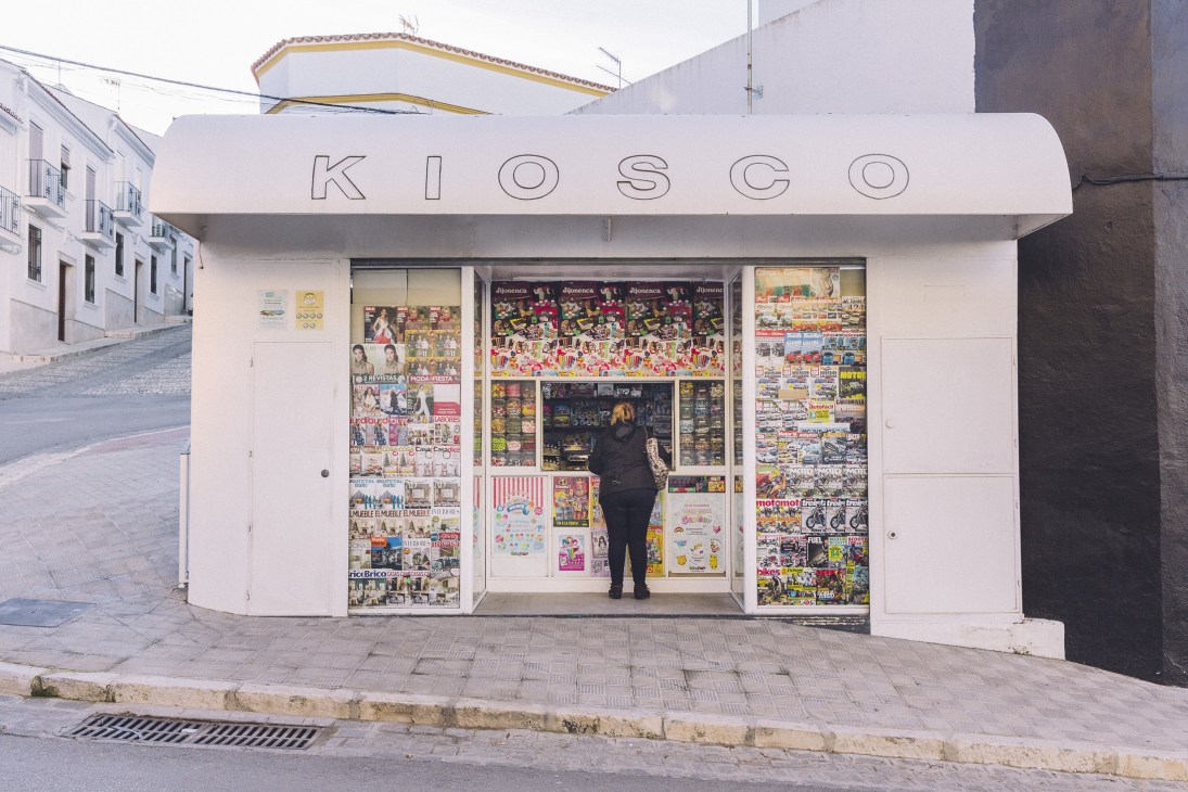 A white kiosk storefront with 'KIOSCO' signage displays newspapers and confectionery products in Estepa.