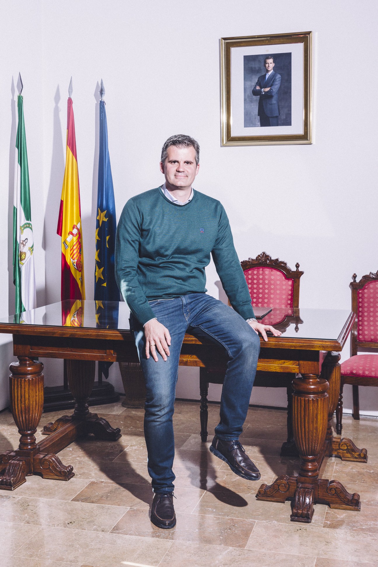 Man sitting on wooden bench with Spanish flags and framed portrait on wall behind him.