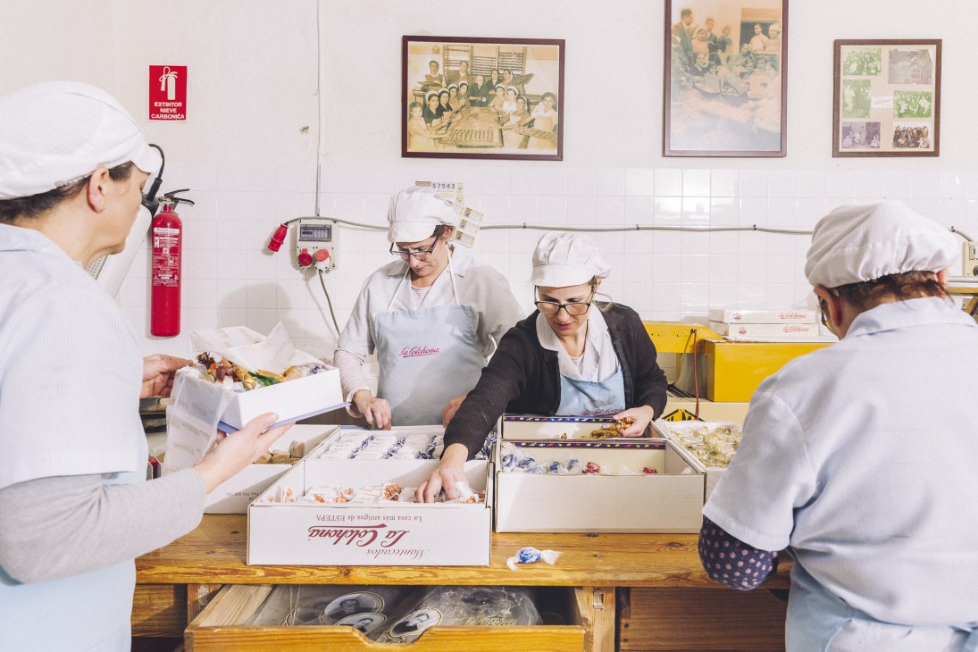 Workers in white uniforms and hairnets hand-wrapping mantecado sweets at La Colchona factory in Estepa, Spain.