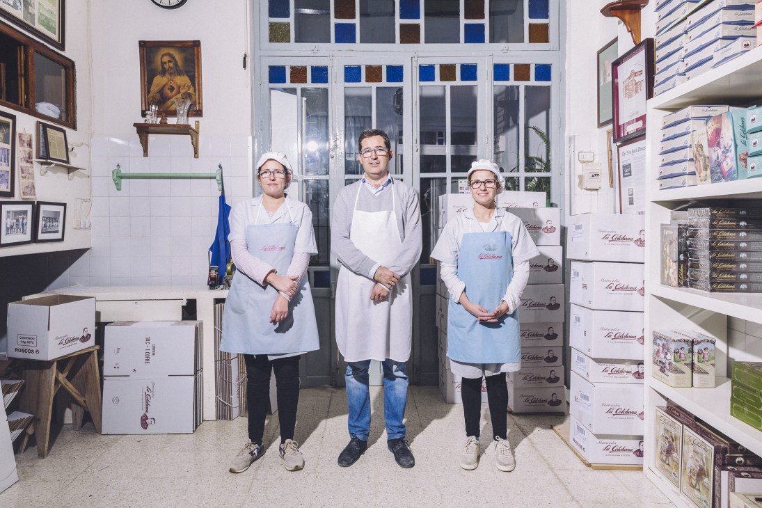 Three workers in white aprons and caps stand in a confectionery workshop surrounded by packaged goods and shelving.