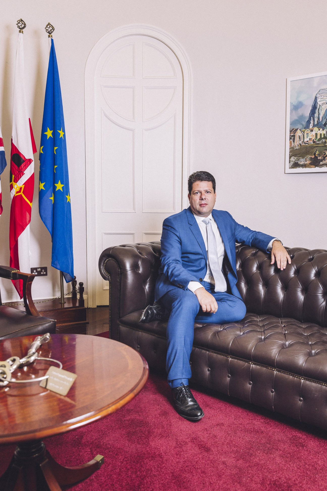 Chief minister Fabian Picardo seated on a leather sofa in an official government office with British and EU flags behind him.