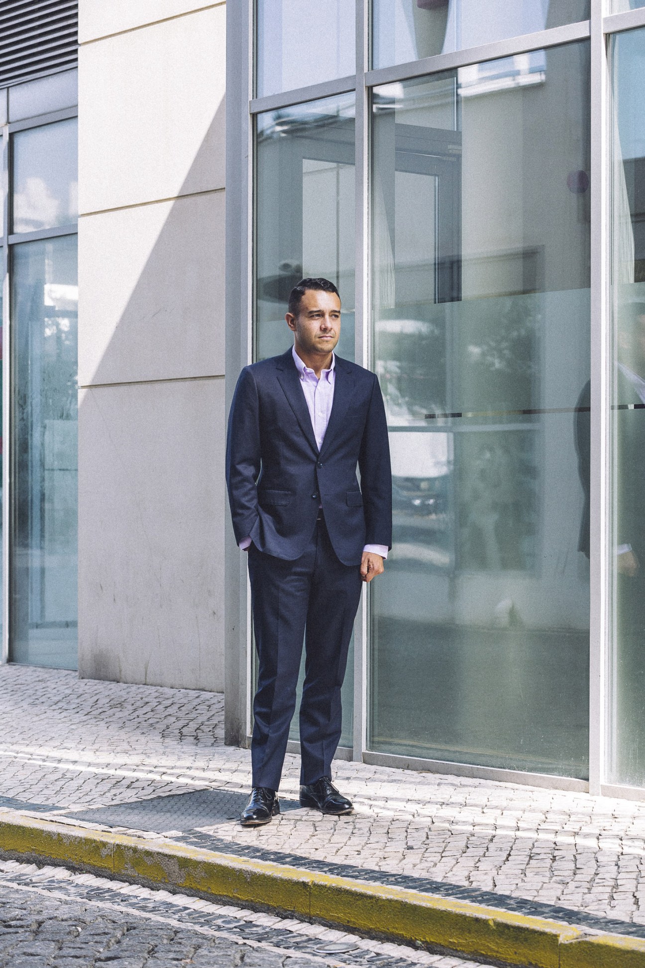 Man in navy blazer and light pink shirt standing outside modern building with glass windows.