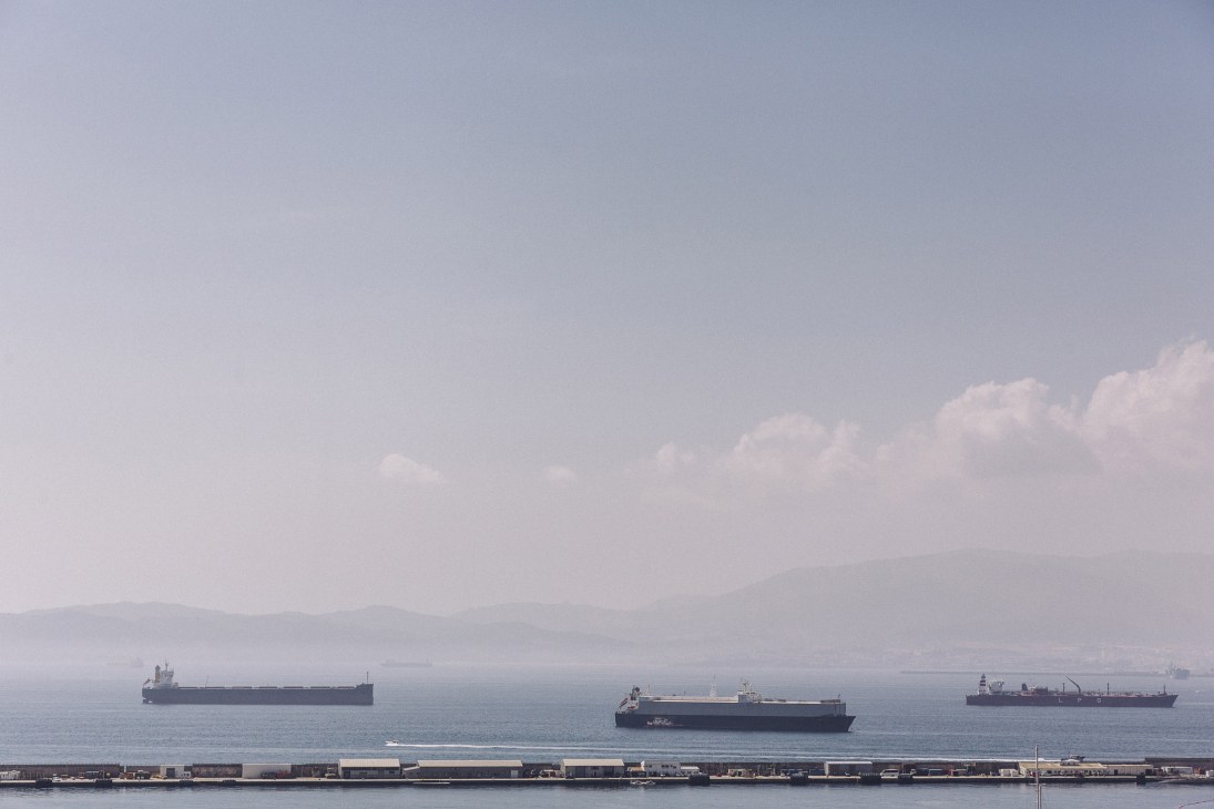 Three cargo ships anchored in waters off Gibraltar with mountains visible across the strait.