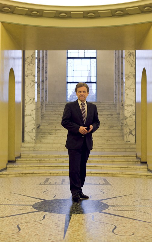 Finland's minister of economic affairs Mauri Pekkarinen standing in a modern government building corridor with yellow walls and marble columns.