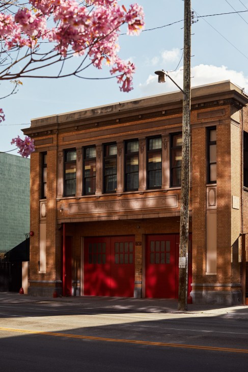 Historic red brick firehouse building with large red doors and flowering pink tree branches in foreground.