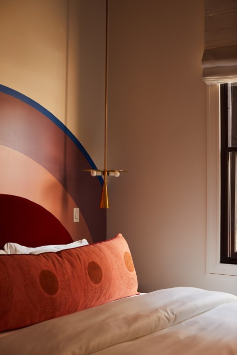 Modern hotel bedroom with coral patterned pillows, pendant light fixture, and curved wall art in warm tones.