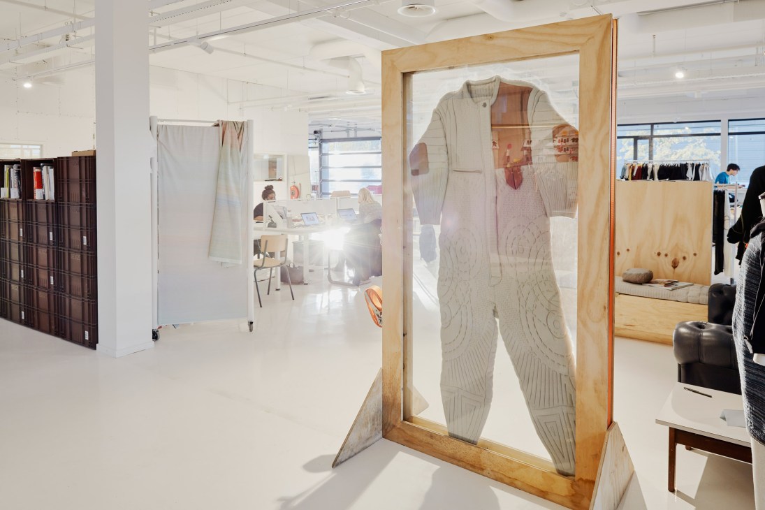 Cream-colored Byborre spacesuit with terracotta panels displayed on wooden frame in textile studio.