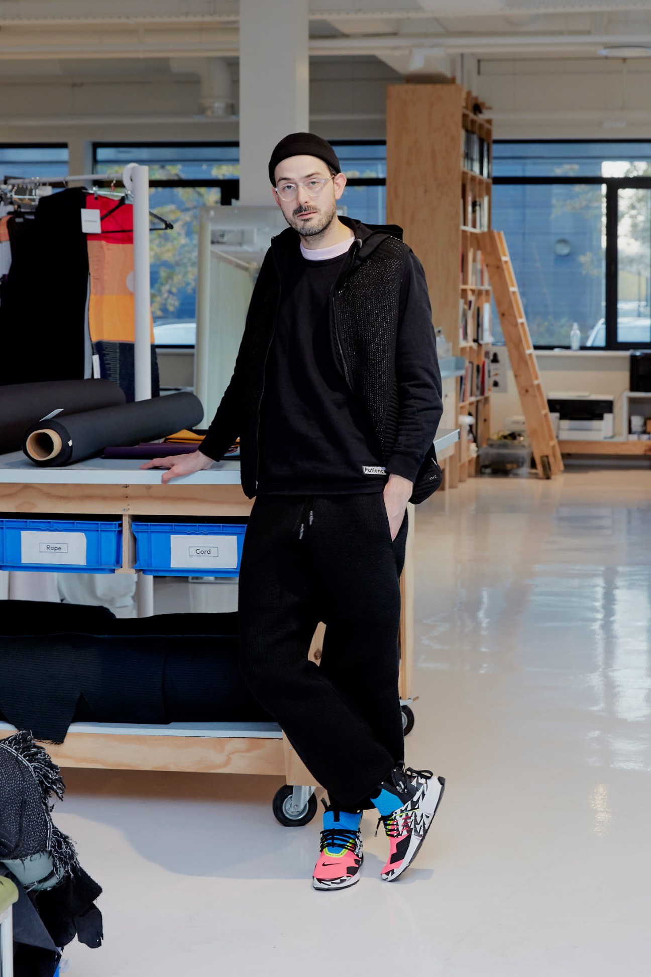 Designer Samuel de Goede stands in a bright studio space with textile materials and knitting equipment visible in the background.