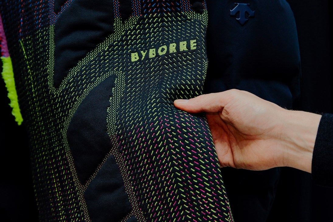 Hand touching a black knitted fabric with neon yellow and pink chevron pattern and Byborre branding.