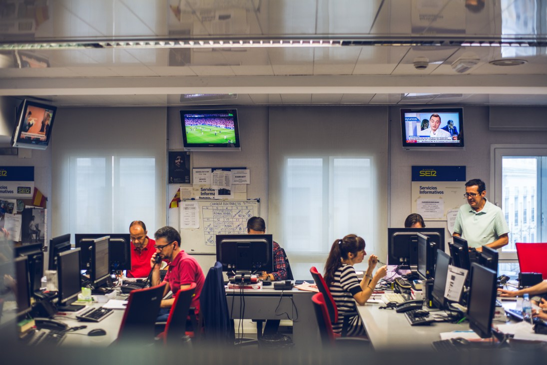 Staff working at desks in Cadena SER's Madrid office with multiple computer monitors and TV screens above.