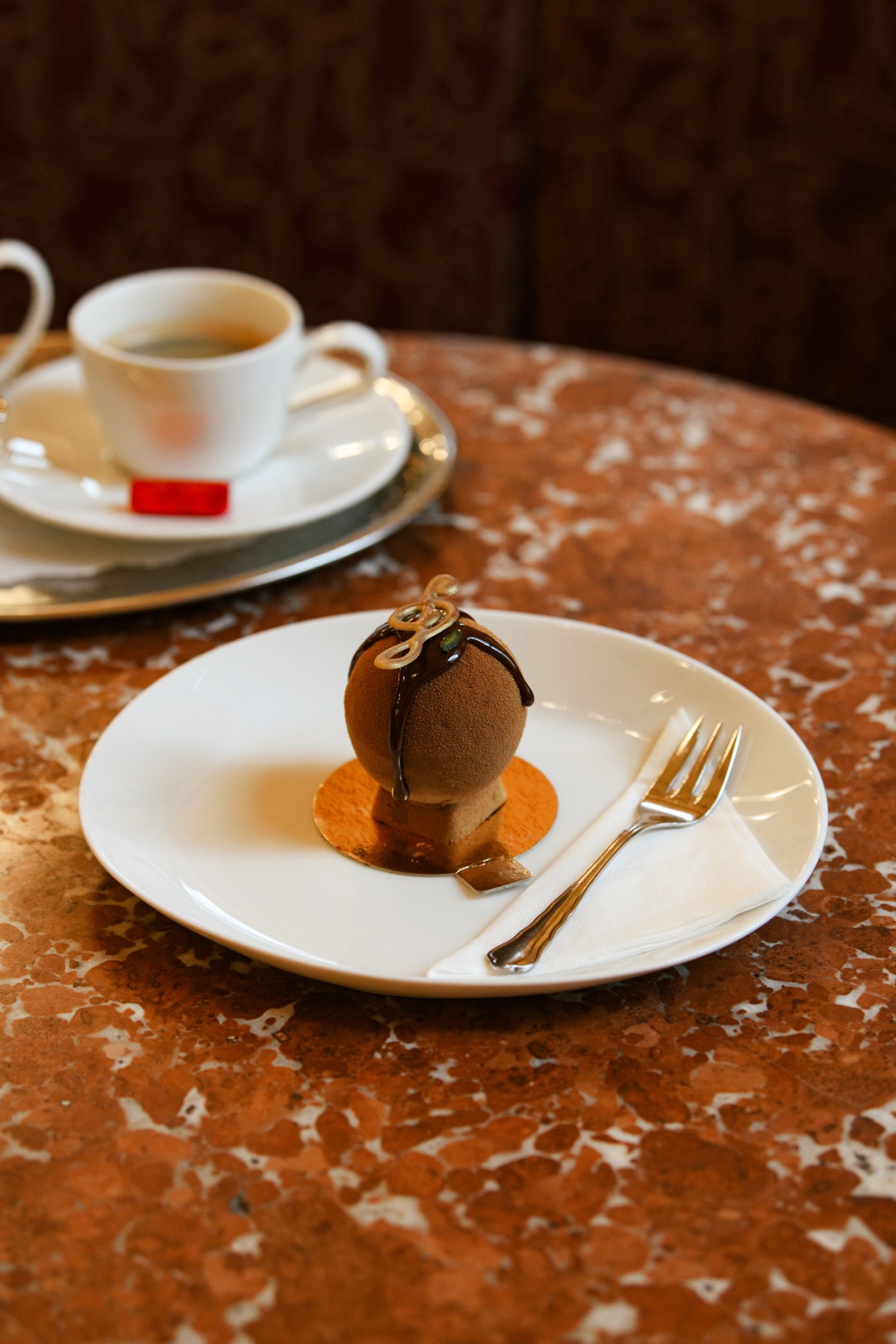 Elegant chocolate dessert with dark glaze and decorative curl on white plate with gold fork beside coffee cup