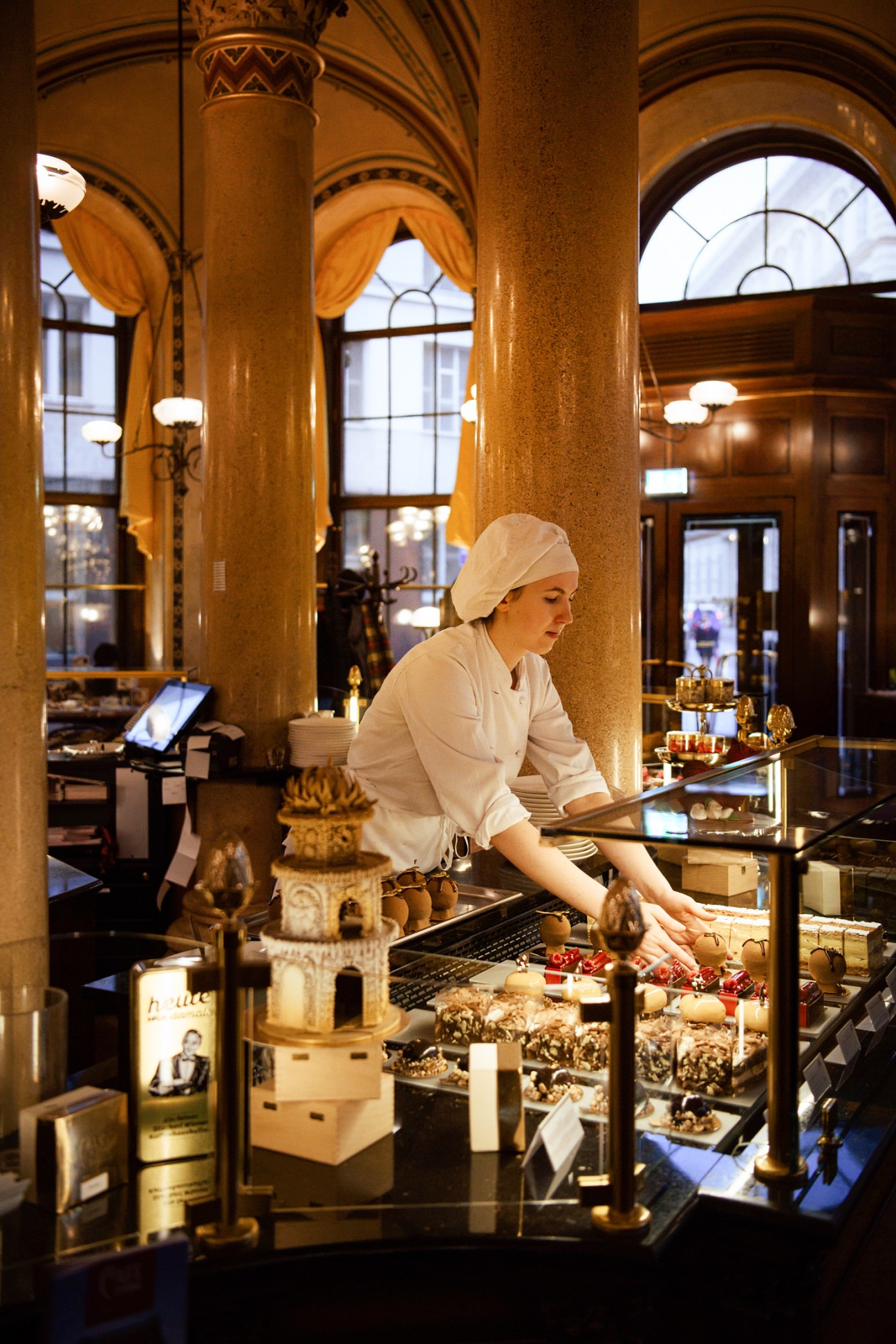 A pastry chef in white uniform and hat arranges desserts in an ornate display case at Café Central