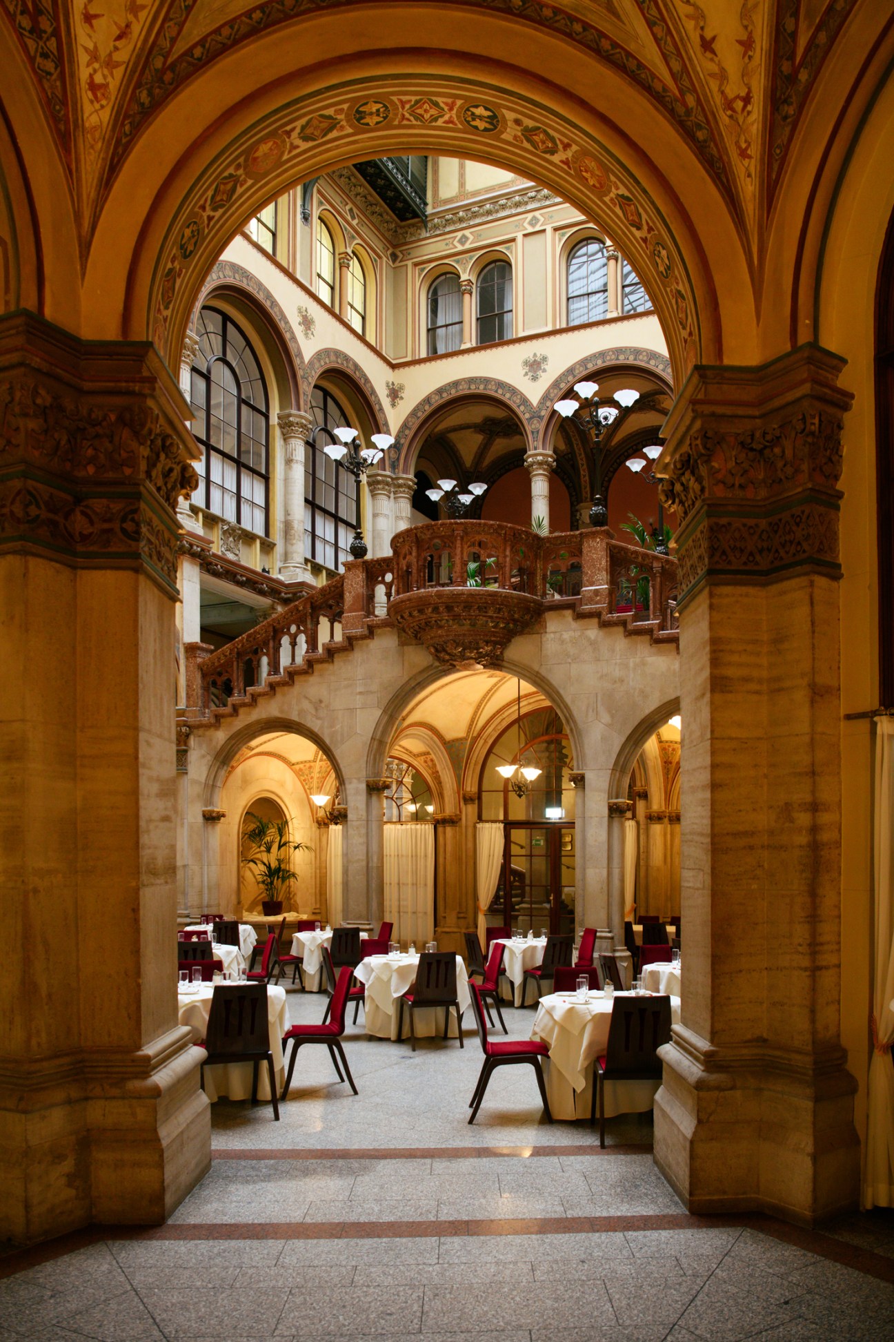 Ornate restaurant dining room with arched stone columns, decorative ceiling, and red chairs at white-clothed tables
