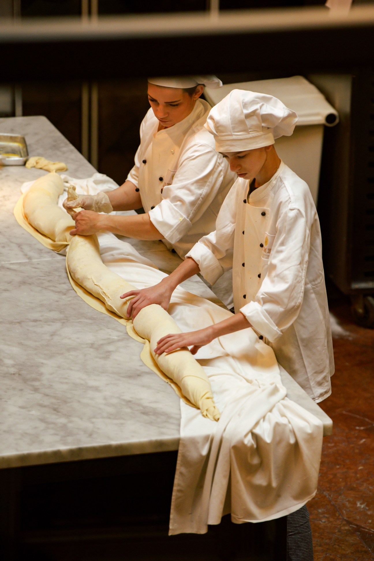 Two chefs in white uniforms and hats working with long strips of pastry dough in Demel's kitchen